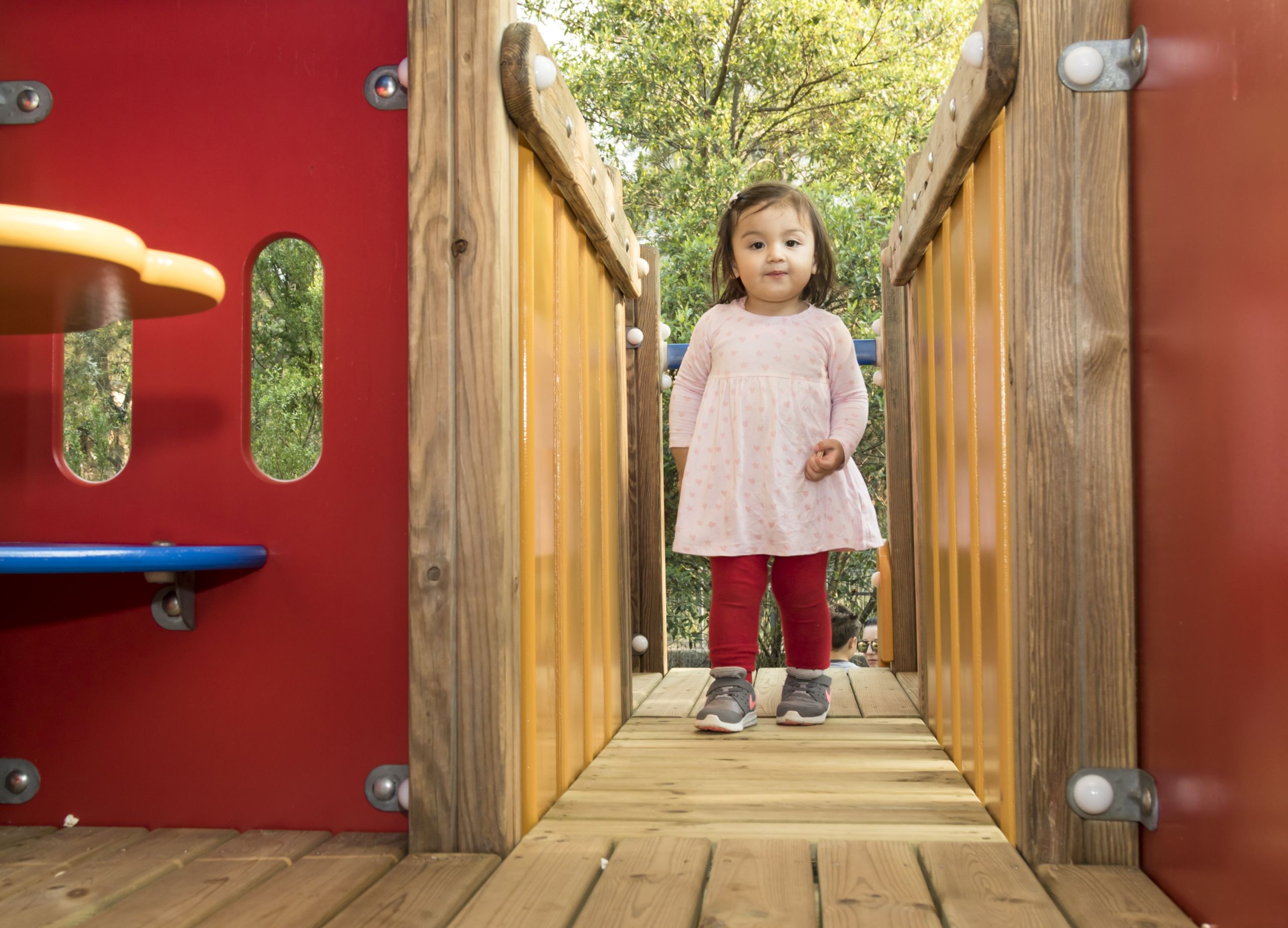 A young girl is walking across an imaginative play bridge. She is smiling.