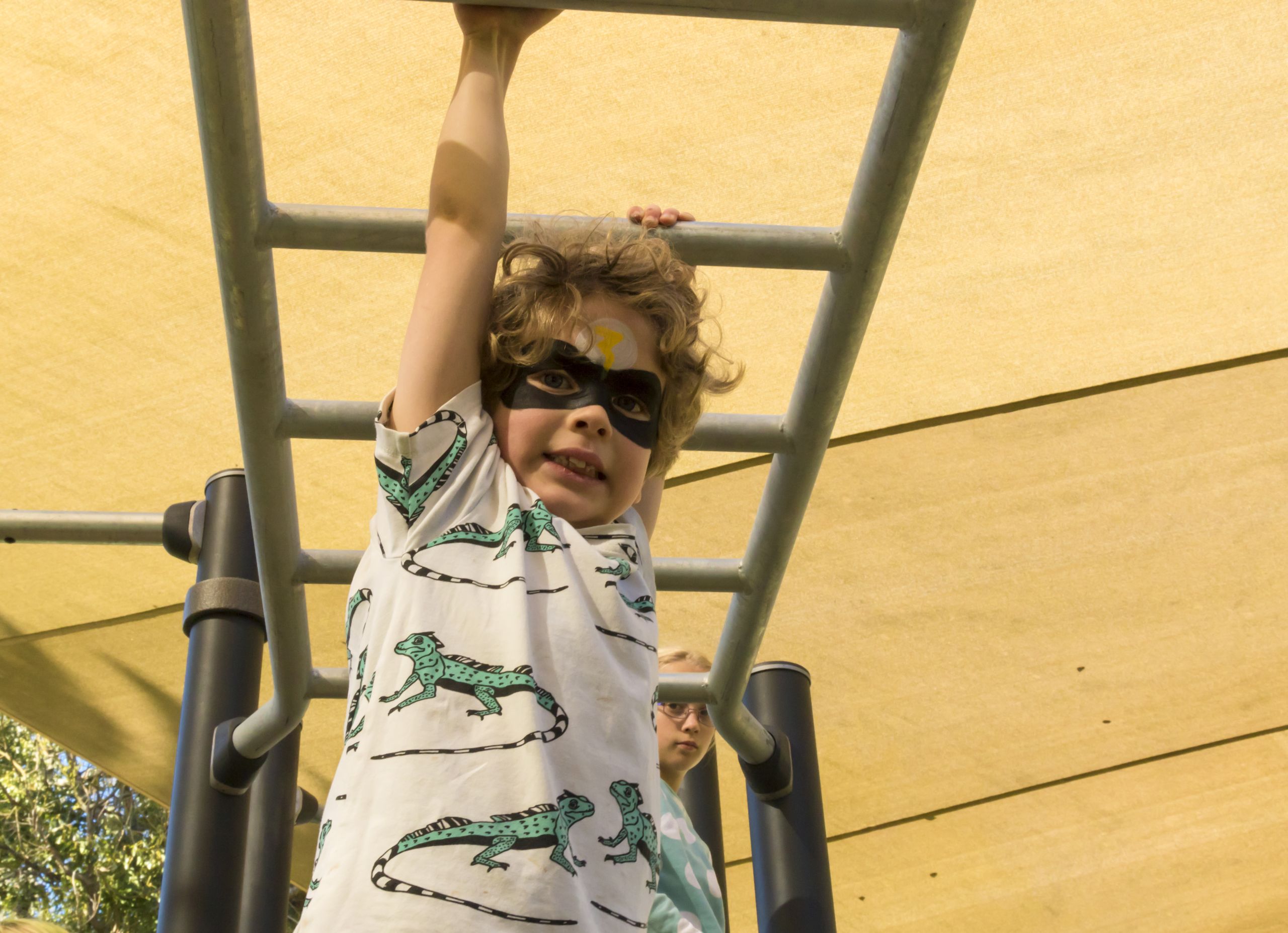 A child wearing a black eye mask is swinging on the monkey bars. He is smiling.