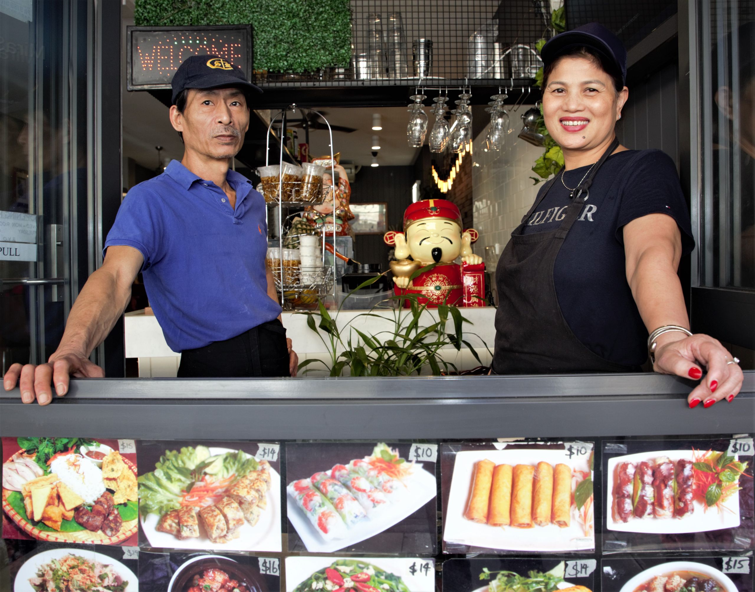 Two staff members stand in the window of a Vietnamese restaurant on Illawarra Road, Marrickville. The menu is presenting in pictures on the shop front.